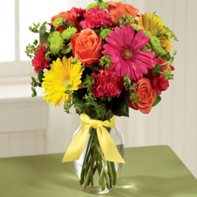 Celebrating life with colorful blooms that inspire and delight, this flower bouquet is ready to create a happy moment for your recipient that they will never forget. Orange roses, hot pink gerbera daisies, yellow gerbera daisies, hot pink carnations, green button poms, bupleurum, and lush greens mingle together to create a sunlit display while seated in a classic clear glass vase tied at the neck with a yellow satin ribbon for a sweet effect.